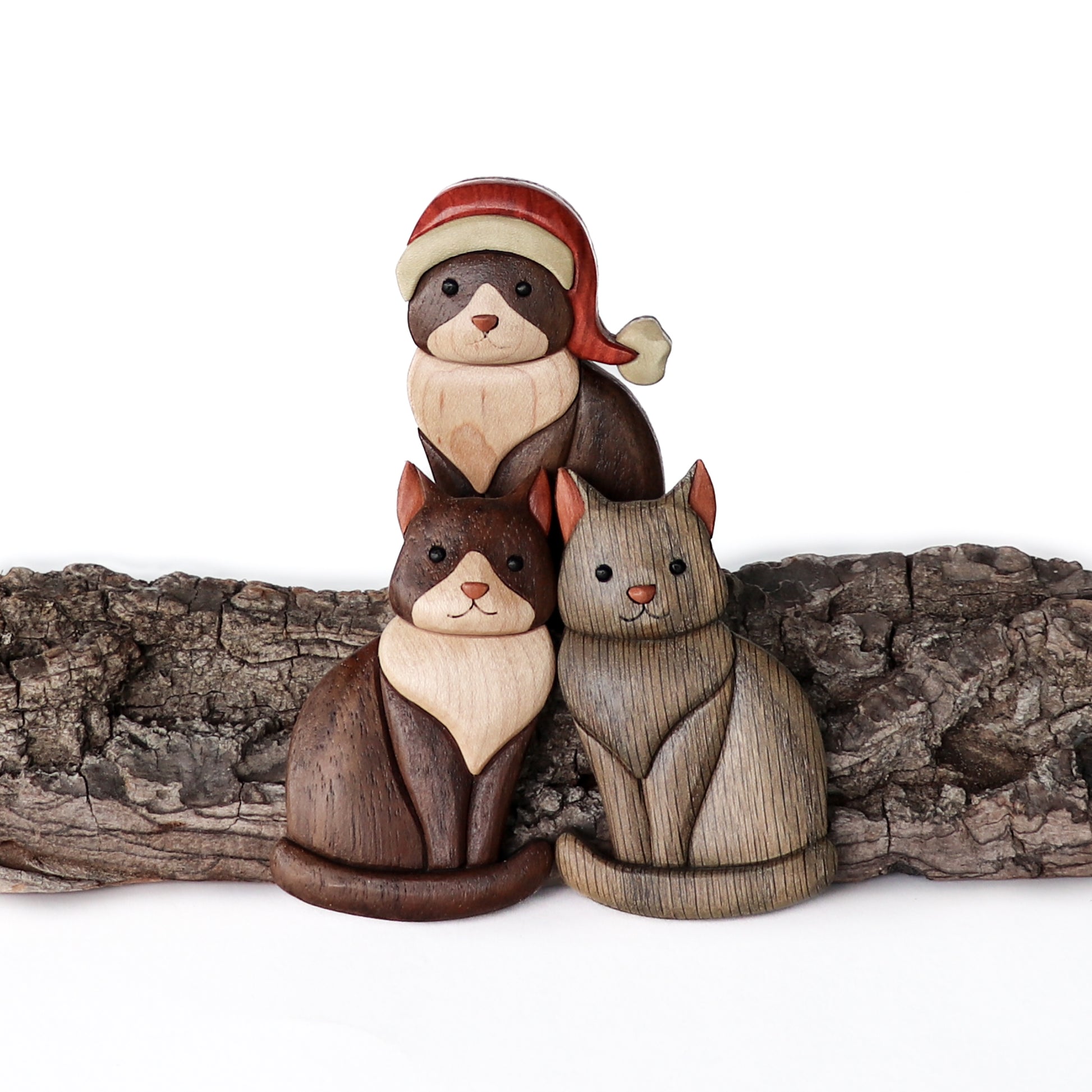 Three different types of cats sitting variations of wood carved intarsia ornaments or magnets with bark.  Two wooden black and white tuxedo cats where one tuxedo cat is wearing a santa hat and one wooden gray cat.