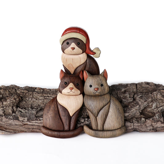 Three different types of cats sitting variations of wood carved intarsia ornaments or magnets with bark.  Two wooden black and white tuxedo cats where one tuxedo cat is wearing a santa hat and one wooden gray cat.