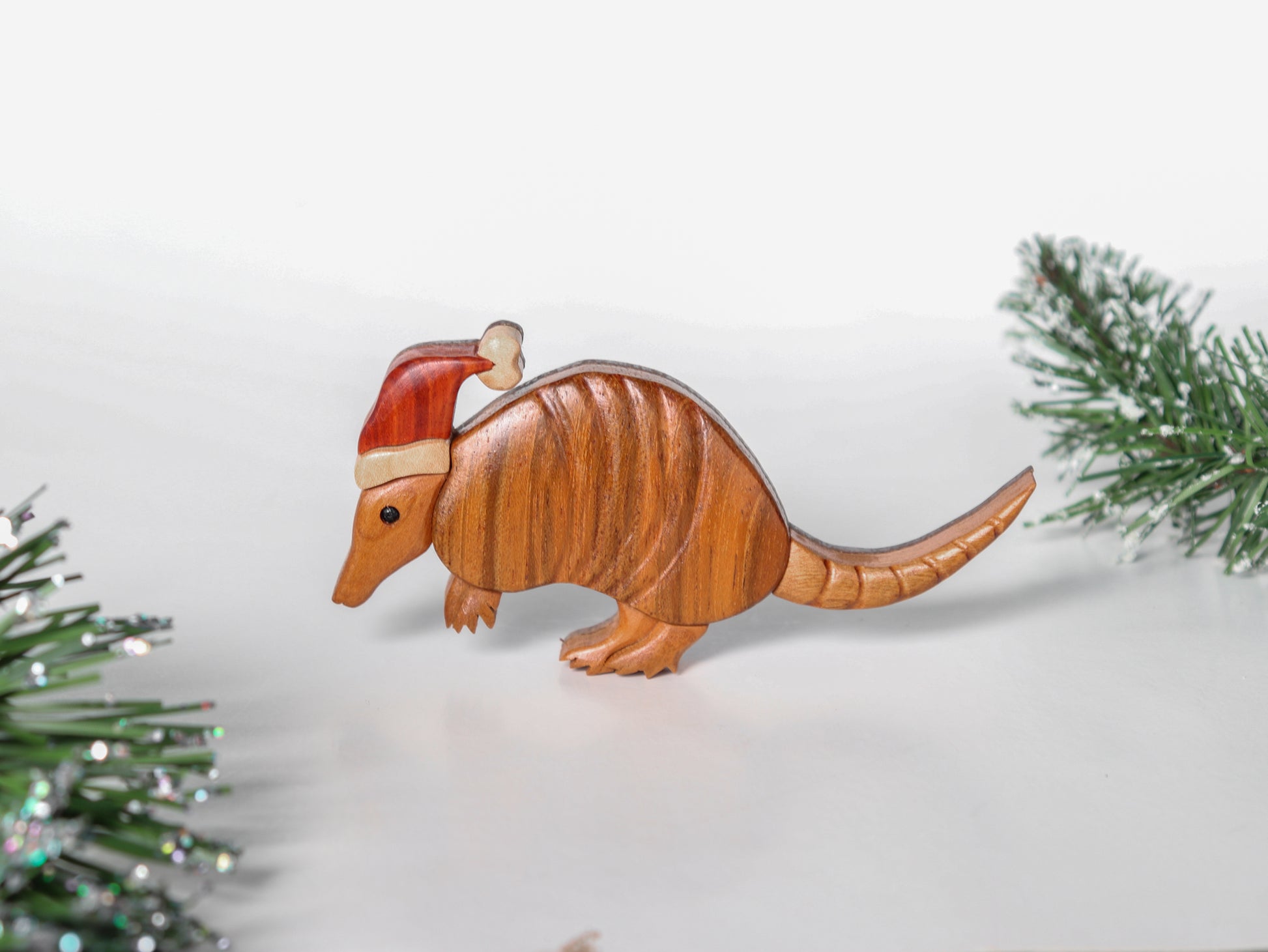 wood carving of an armadillo wearing a chritmas hat made from redheart and holly, surrounded by conifer leaves and branches.