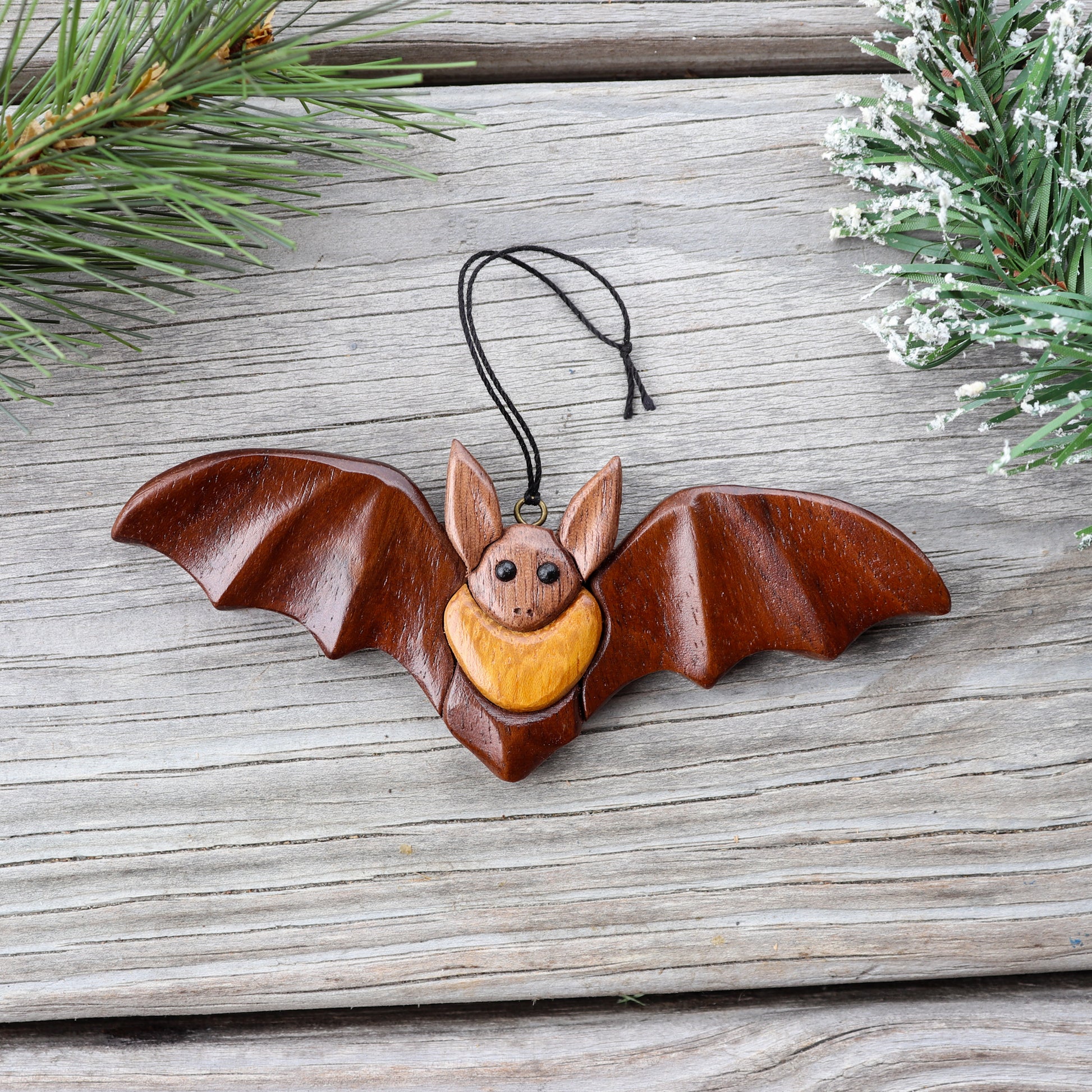 wooden bat ornament intarsia laying down on a plank, surrounded by pine branches, showing the carving details of the folds of the wings outstretched.