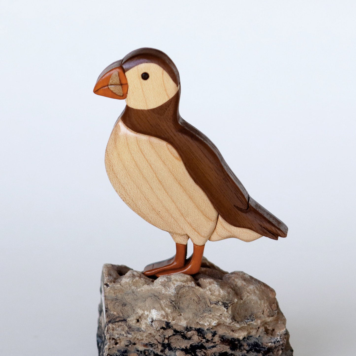 wood carving of an atlantic puffin using the intarsia style woodworking made using maple, peruvian walnut, chakte viga, standing on a rock, really showing the woodgrain of the maple.