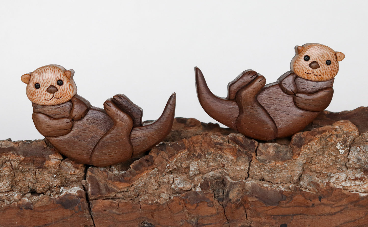 Mirror image of two wooden otter intarsia made from peruvian walnut and sycamore, laying on their back on black walnut bark.