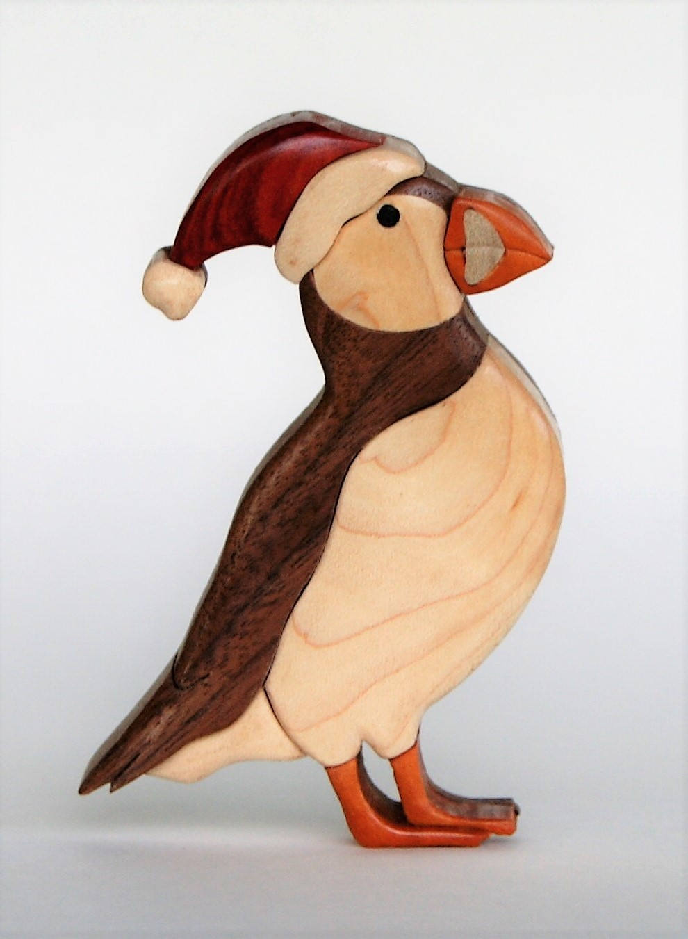wood carving of a christmas puffin wearing a santa hat made from redheart and maple, standing facing left, showcasing the woodgrain of the maple following the body.