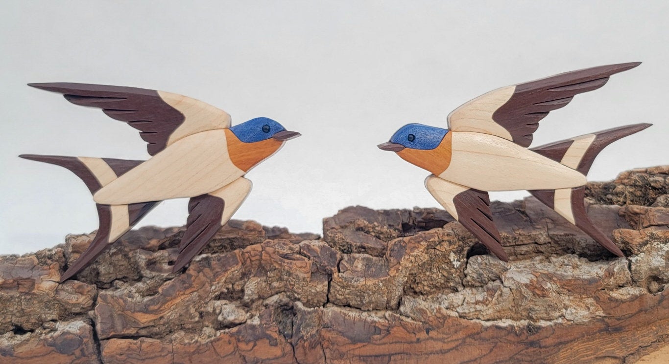 two wooden barn swallow in flight decorations showing the woodgrain of maple with blue dye on the head, facing each other on top of a piece of bark.