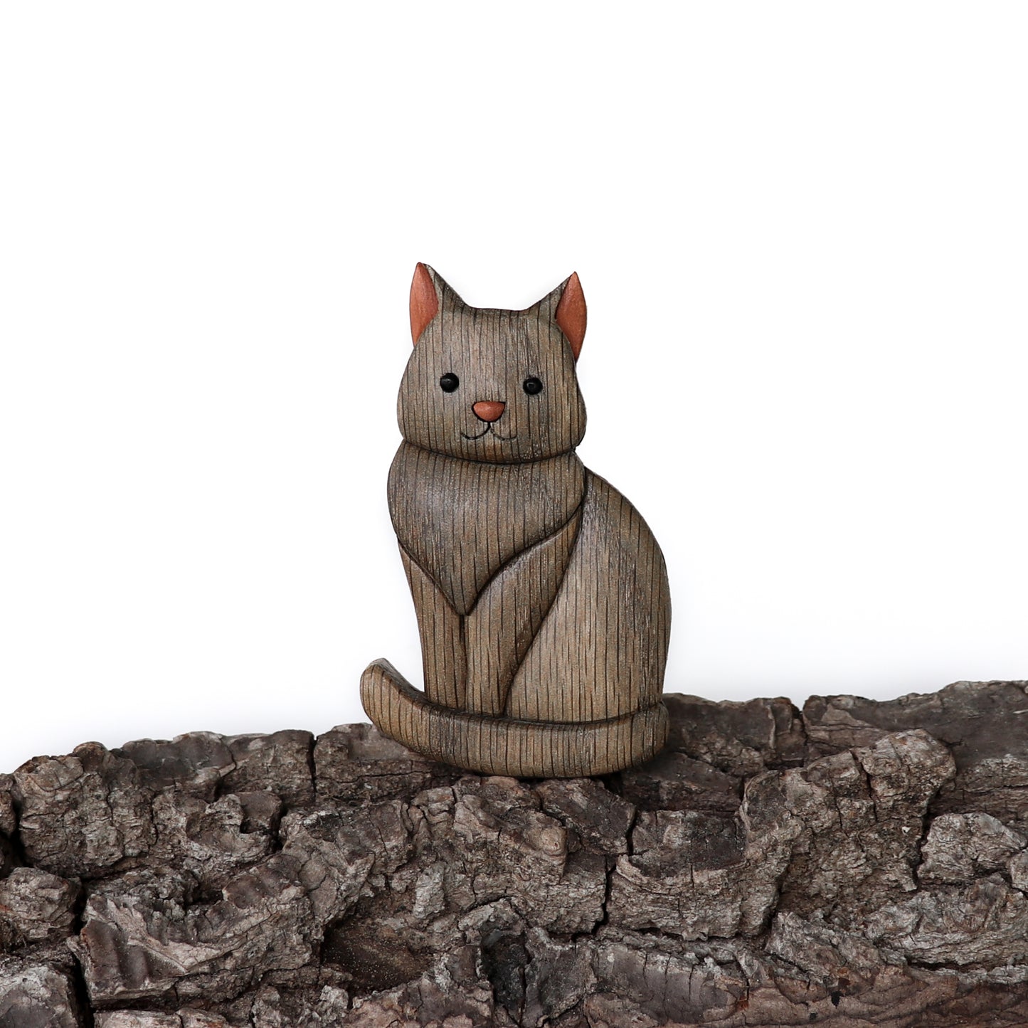 Wood carved art of a gray cat and pink nose and ears sitting on top of bark and with a white background