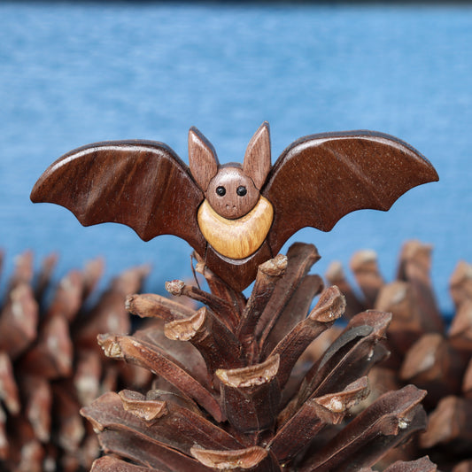 wood carving of a bat with wings outstretched and pointy ears, made from peruvian walnut, black walnut, and iroko with wood burned dowel eyes, perched on a long leaf pine cone with a blurred blue background.